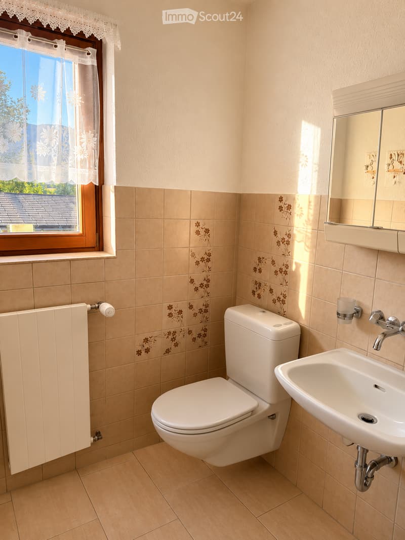 Bathroom with beige tiles, a toilet, a sink, a mirror, and a radiator. The window has lace curtains and offers a view of the roof.