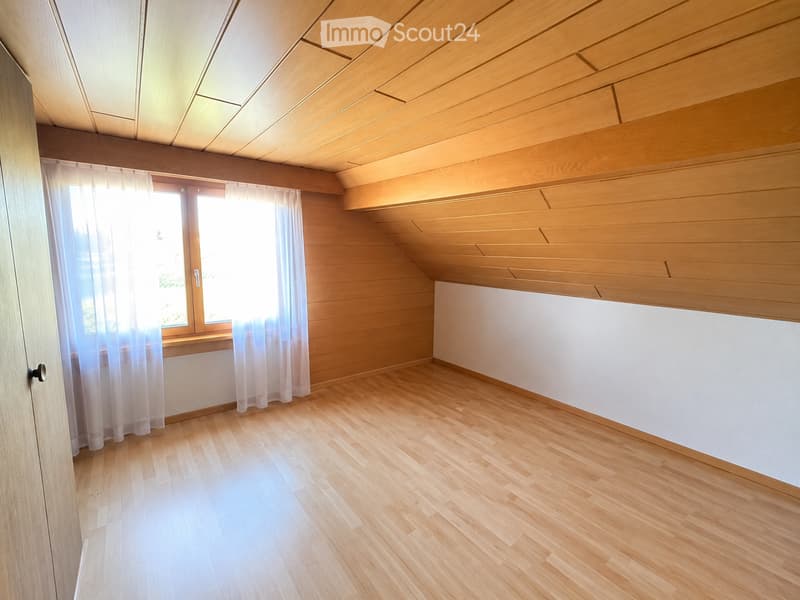 Empty room with wooden flooring, white walls, large window with sheer curtains, and wooden ceiling beams.