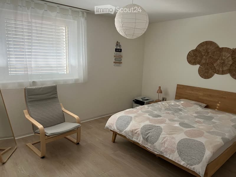 Bright bedroom with wooden flooring, a large bed with a stone-patterned duvet, a wooden chair, a small table lamp, and decorative wall art featuring circular wooden designs.