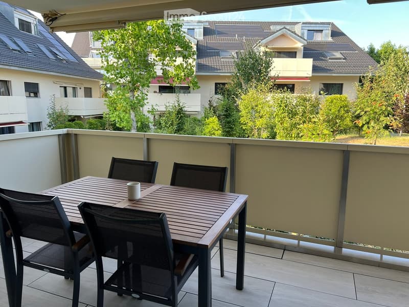 Balcony with a wooden table, four black chairs, and a white cup, overlooking a residential area with trees and houses.