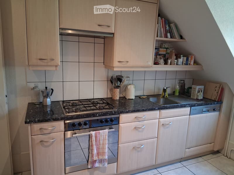 Compact kitchen with light wooden cabinets, gas stove, oven, dishwasher, and countertop with various kitchen items including a kettle, cup, and coffee maker.