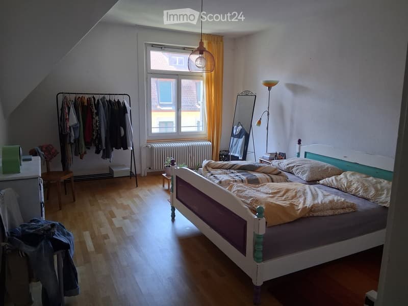 Cozy attic bedroom with wooden flooring, a white bed with patterned bedding, a large window with yellow curtains, a clothing rack, a small table, and a mirror.