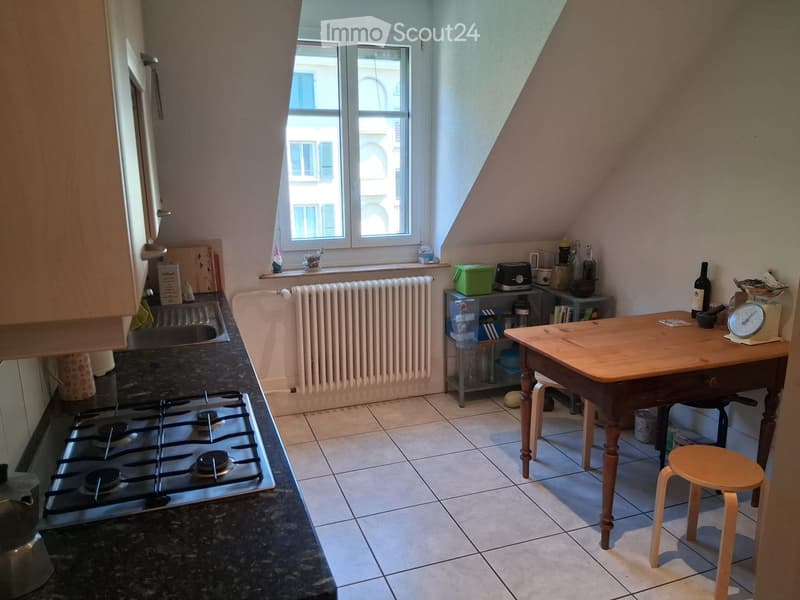 A compact kitchen with a gas stove, sink, wooden table, and small stool. The space features tiled flooring, a radiator, and a window with a view of the street.