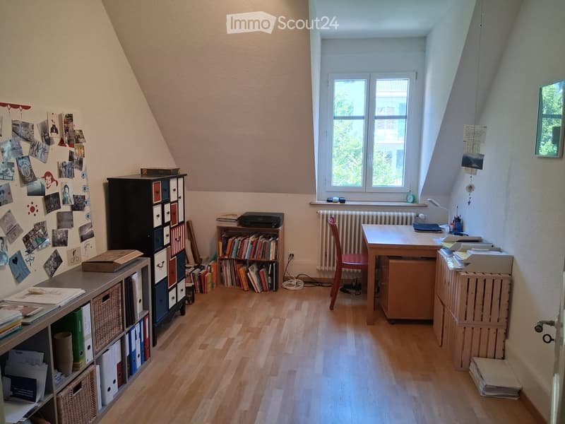 A well-lit attic room with wooden flooring, large windows, and various furniture including a desk, chair, bookshelf, and cabinet.