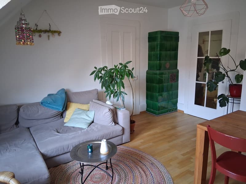 Living room with a grey sectional sofa, coffee table, small round table, red chair, and large green plant. Features wooden flooring, a green tiled stove, and decorative hanging lights.