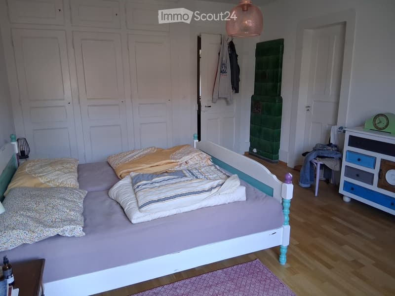 Cozy bedroom with a double bed, colorful bedding, a nightstand with a lamp, a wardrobe, and a small chair near the door.