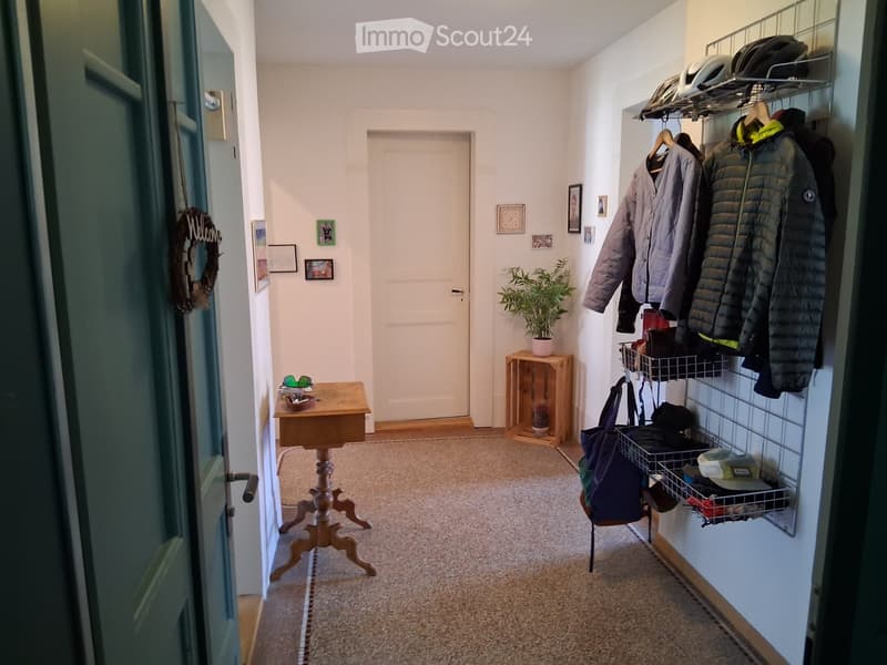 A well-lit hallway with a small wooden table, coat rack, and potted plant. The hallway features a carpeted floor and several framed pictures on the walls.