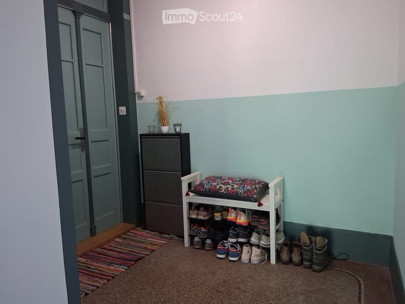 A small entryway with a shoe rack, a small cabinet, and a decorative pillow on top. The walls are painted in two tones, and a colorful rug is placed on the floor.