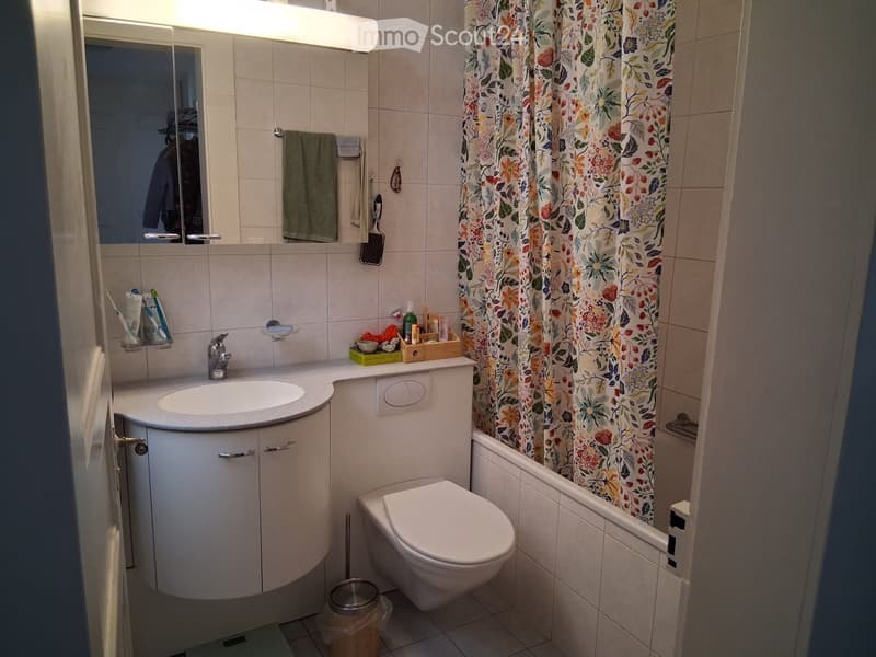 Modern bathroom with a sink, toilet, mirror, and a shower area with a floral curtain. Includes a towel, toothbrush, toothpaste, and a small trash can.