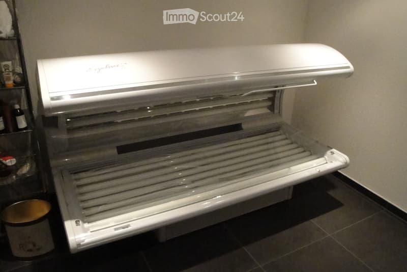 A white refrigerator with multiple shelves and a top-hinged door, placed in a room with tiled flooring and a shelf unit in the background.