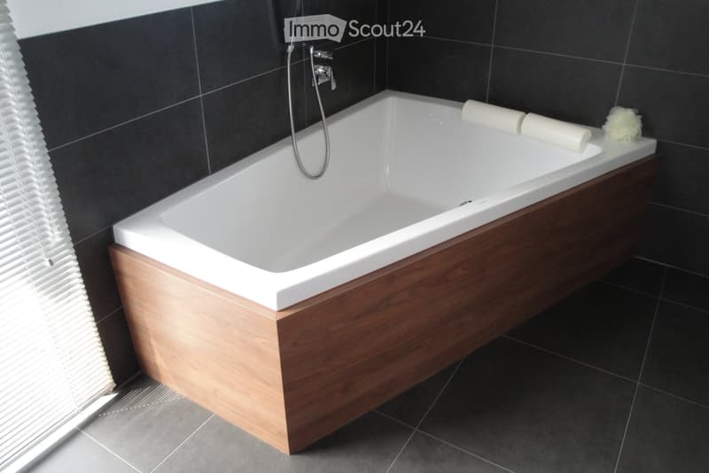 Modern bathroom with a white bathtub, wooden tray, and towels placed on the edge. Dark tiled walls and floor, with a window with blinds on the left side.