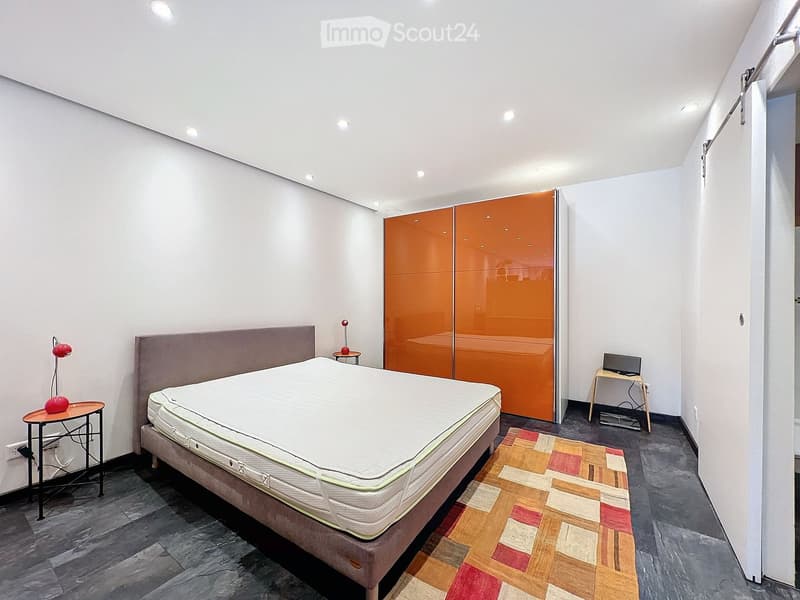 Modern bedroom with a large bed, orange wardrobe, small table with a lamp, and a small desk with a screen.