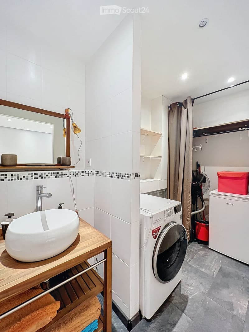 Modern bathroom with a white sink, wooden shelf, and a washing machine in the adjacent laundry area.
