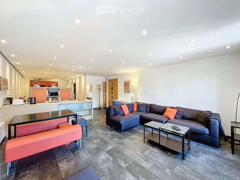 Modern living room with a large sectional sofa, coffee tables, a bench, and a small kitchen area with appliances.