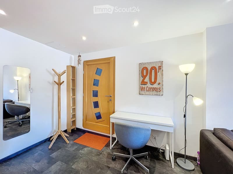 A modern, well-lit room with a desk, chair, sofa, coat rack, shelf, and a mirror. The room features a wooden door with blue square panels and a poster on the wall.
