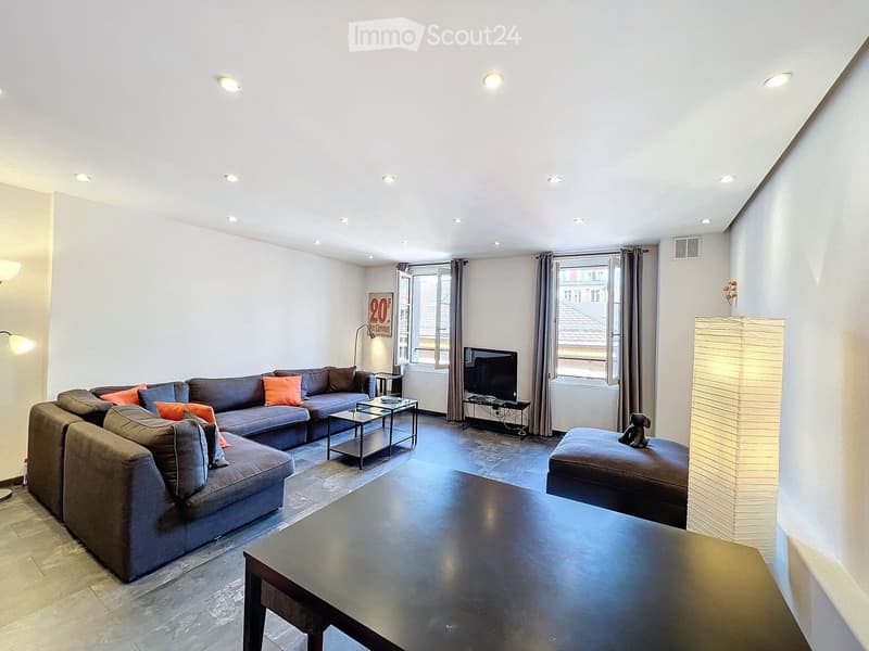Modern living room with grey sectional sofa, coffee table, ottoman, and a television on a stand. The room features large windows with curtains and recessed ceiling lighting.