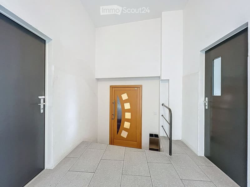 Modern hallway with tiled floor, white walls, and three doors: a wooden door with square panels, a dark grey door, and another dark grey door with a small mirror. A metal handrail is visible on the right side.