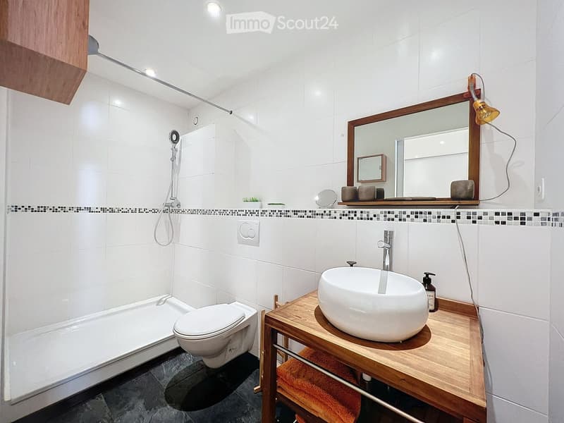 Modern bathroom with white tiles, a bathtub, toilet, sink, and a large mirror with wooden frame. Includes a wooden vanity with a sink, towel, and decorative items on the counter.