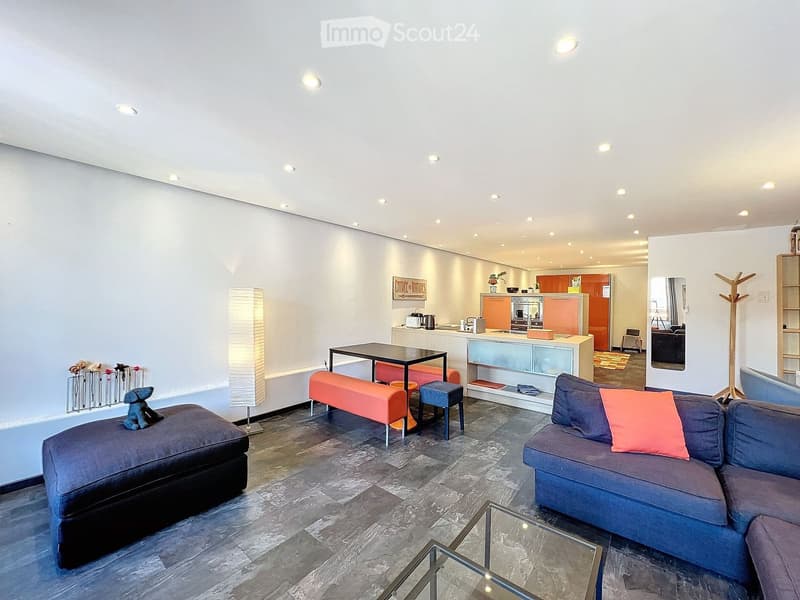Modern living room with a grey sectional sofa, glass coffee table, bench seating, and a small table with stools. The space features a sleek design with a mix of dark and light tones, a tall floor lamp, and a small kitchen area visible in the background.