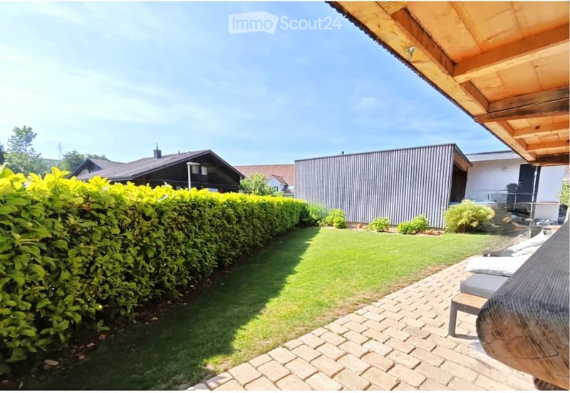 Well-maintained garden with a neatly trimmed hedge, a brick patio, and a lounge chair, surrounded by houses and greenery.