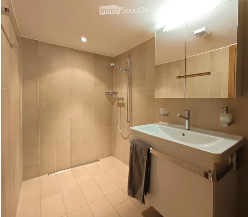 Modern bathroom with beige tiles, a white sink, a mirror cabinet, a shower, and a towel hanging on the side.