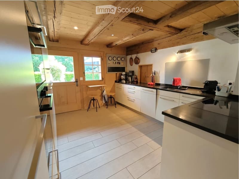 Modern kitchen with white cabinets, black countertops, and stainless steel appliances. Features include a sink, coffee maker, frying pans, and stools near the window.