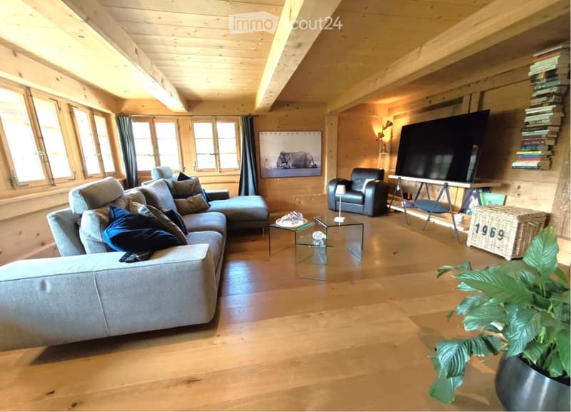 Spacious living room with wooden beams and exposed wooden ceiling, large windows, grey sectional sofa, coffee table, armchair, TV screen, and decorative plant.