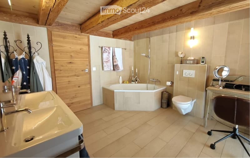 Modern bathroom with wooden beams, a large sink, bathtub, toilet, and a vanity with a mirror. Features include a towel rack with towels, a light fixture, and a chair near the vanity.