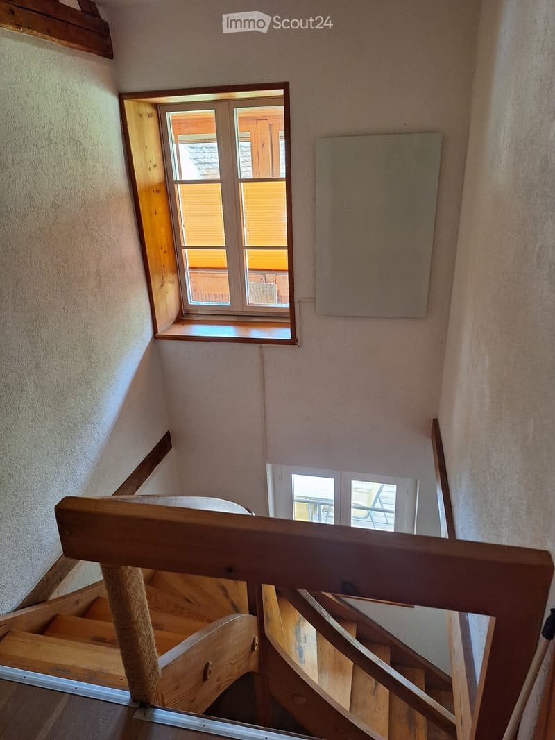 Wooden staircase with a handrail, light-colored walls, and a window overlooking a room with wooden furniture and a view of the roof.