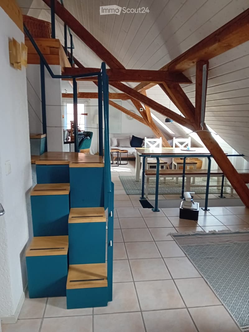 Loft-style living area with exposed wooden beams, modern furniture including a sofa, dining table, chairs, and wooden stools, with a staircase leading upwards.