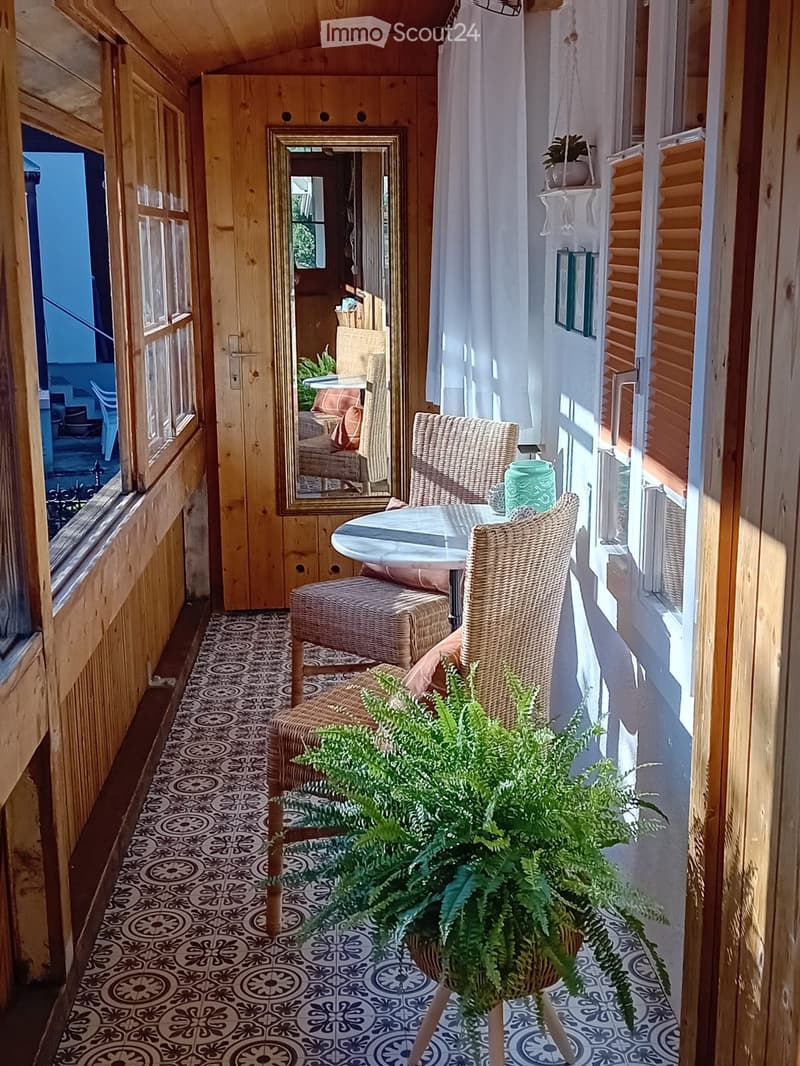 Balcony with wooden walls, patterned tile floor, round table, two wicker chairs, and a potted plant.