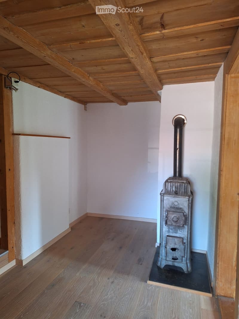 Empty room with wooden floor, white walls, exposed wooden ceiling beams, and a metal wood-burning stove with a chimney pipe.