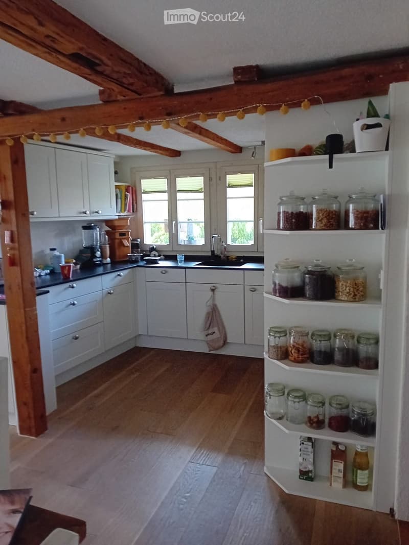 Bright kitchen with white cabinets, wooden floor, and large windows. Features a blender, coffee grinder, sink, and shelves filled with jars and food items.