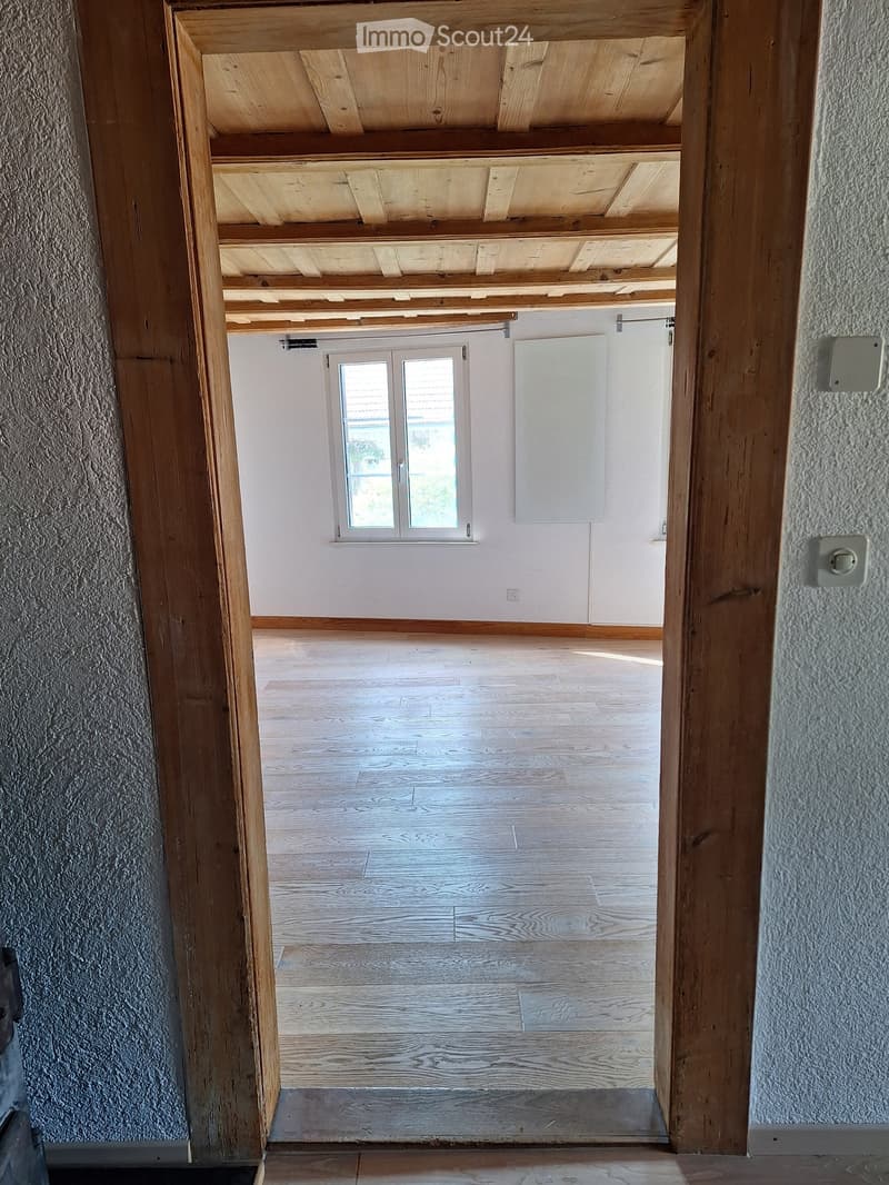 Empty room with wooden floor, white walls, and a window with double glass panels. Exposed wooden beams on the ceiling.