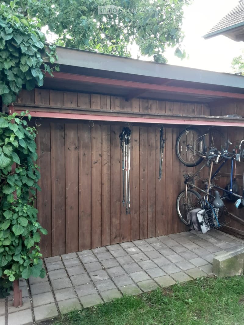 Wooden shed with bicycles and ski poles stored inside, surrounded by greenery and a paved area.