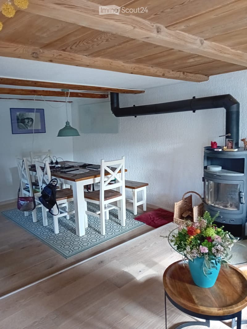 Cozy living and dining area with wooden floors, exposed wooden beams, a dining table with chairs, a small round table with a flower vase, and a wood-burning stove.