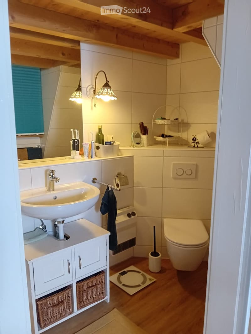 Modern bathroom with a white sink, faucet, mirror, toilet, and cabinet. Features wicker baskets on the cabinet, a towel hanging on a rack, and decorative lighting.