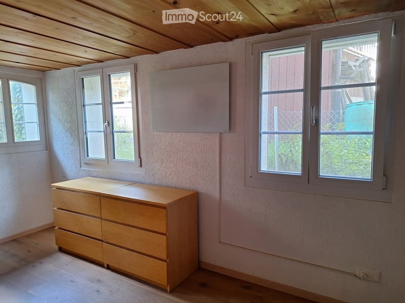 Empty room with light wooden floor, light pink walls, and a chest of drawers near the window. The room has multiple windows with white frames and a visible electrical outlet on the wall.