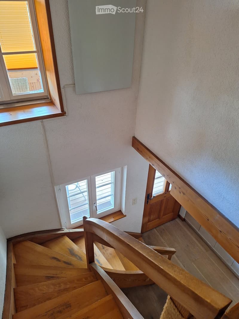 Wooden staircase with a wooden handrail, light-colored walls, and windows with blinds. The staircase leads to a landing area with a door and a small window.