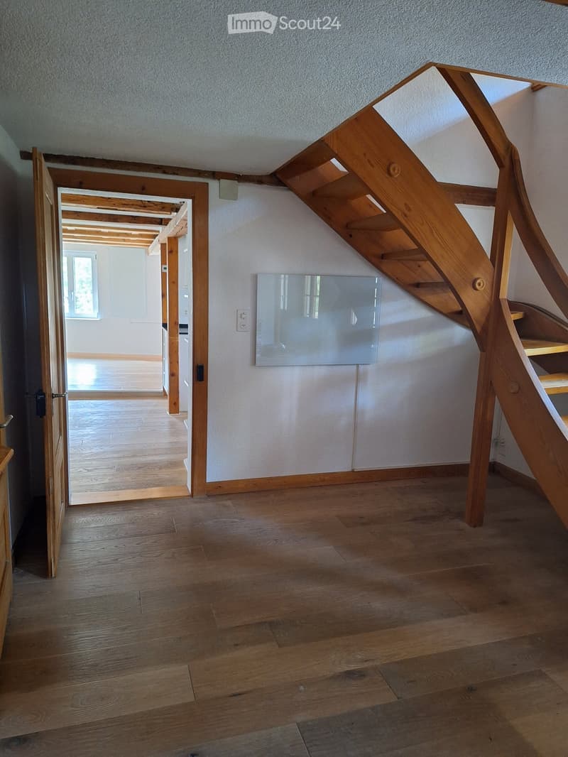 Wooden staircase with glass panel, open doorway leading to another room, wooden flooring, white walls, visible electrical outlet.