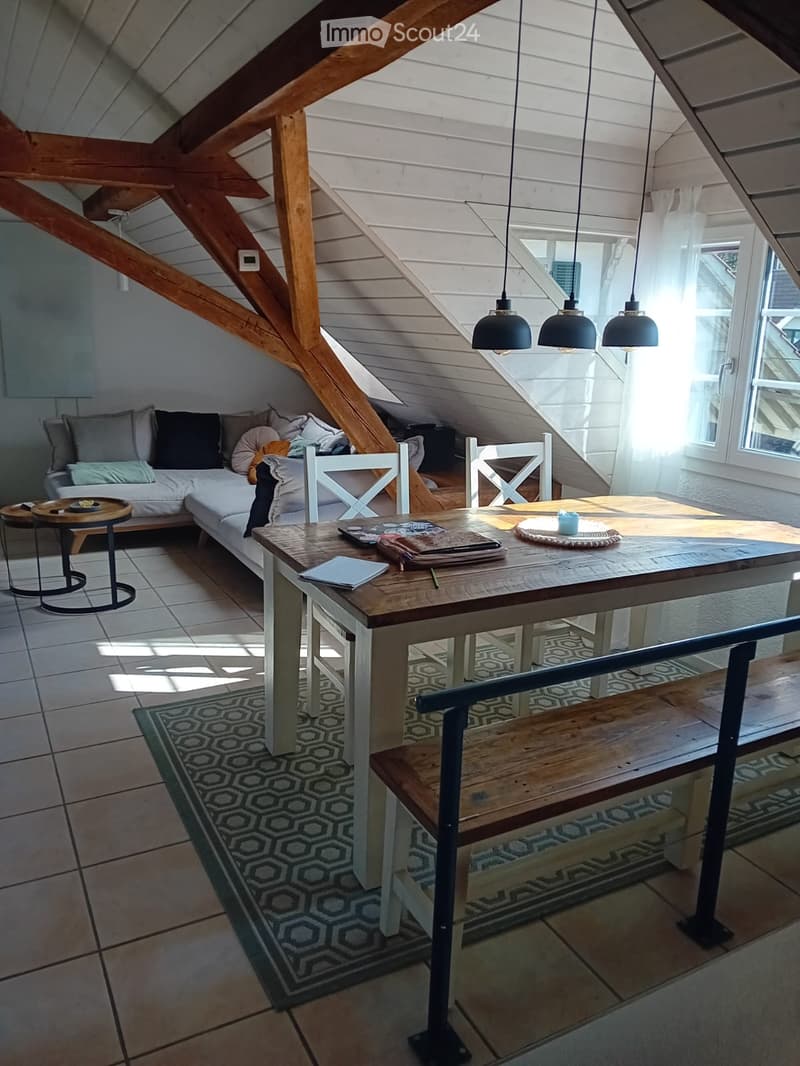 Loft-style room with exposed wooden beams, white walls, and large windows. Features a white sectional sofa, wooden dining table with chairs, pendant lights, and a bench.