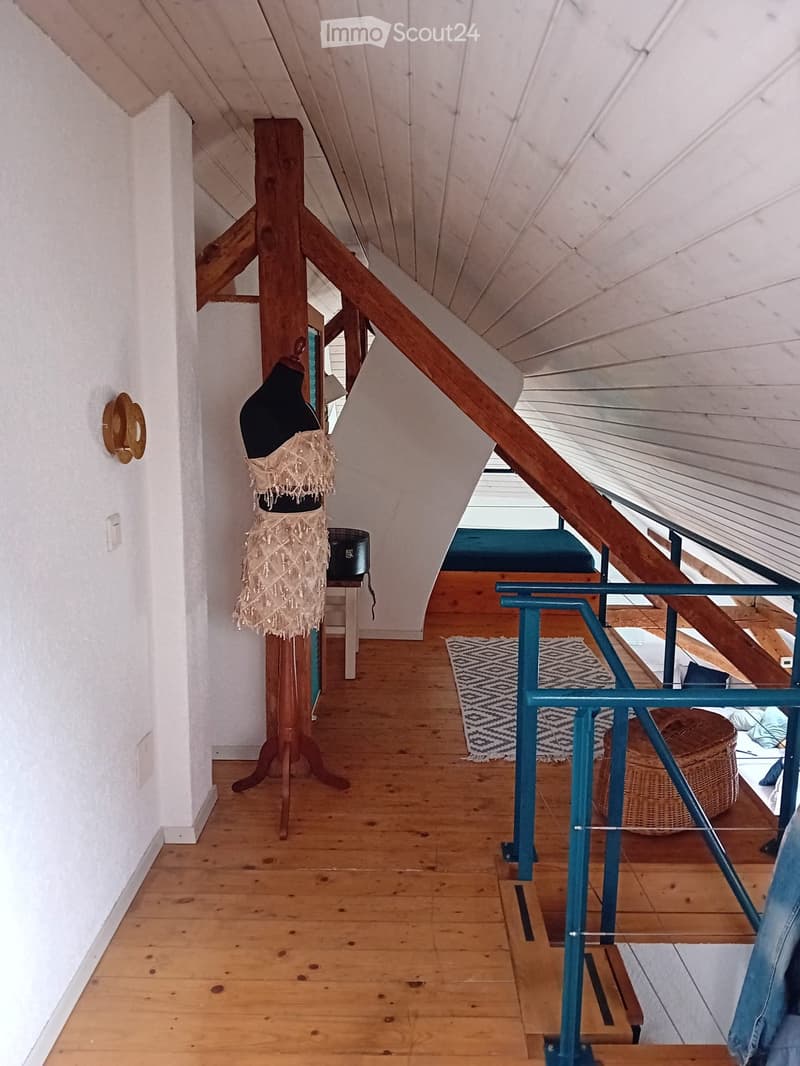 A wooden staircase leads up to a loft area with exposed wooden beams and a slanted ceiling. The hallway features a mannequin, a small table, and a patterned rug on the floor.