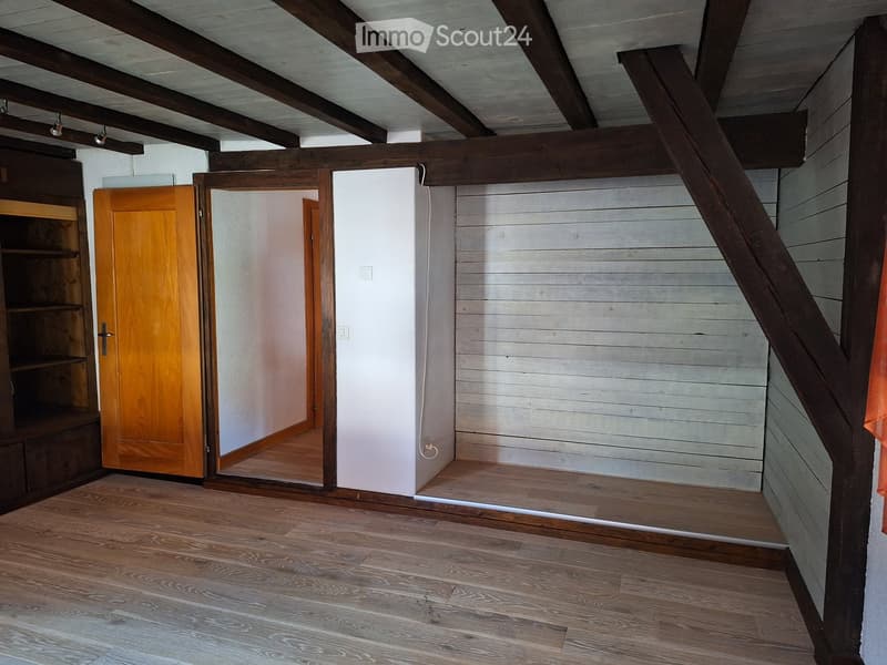 Empty room with wooden flooring, exposed wooden beams on the ceiling, and a wooden door leading to another space.