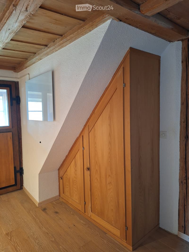 An unfurnished room with wooden flooring, exposed wooden beams on the ceiling, a wooden closet, and a small mirror on the wall.
