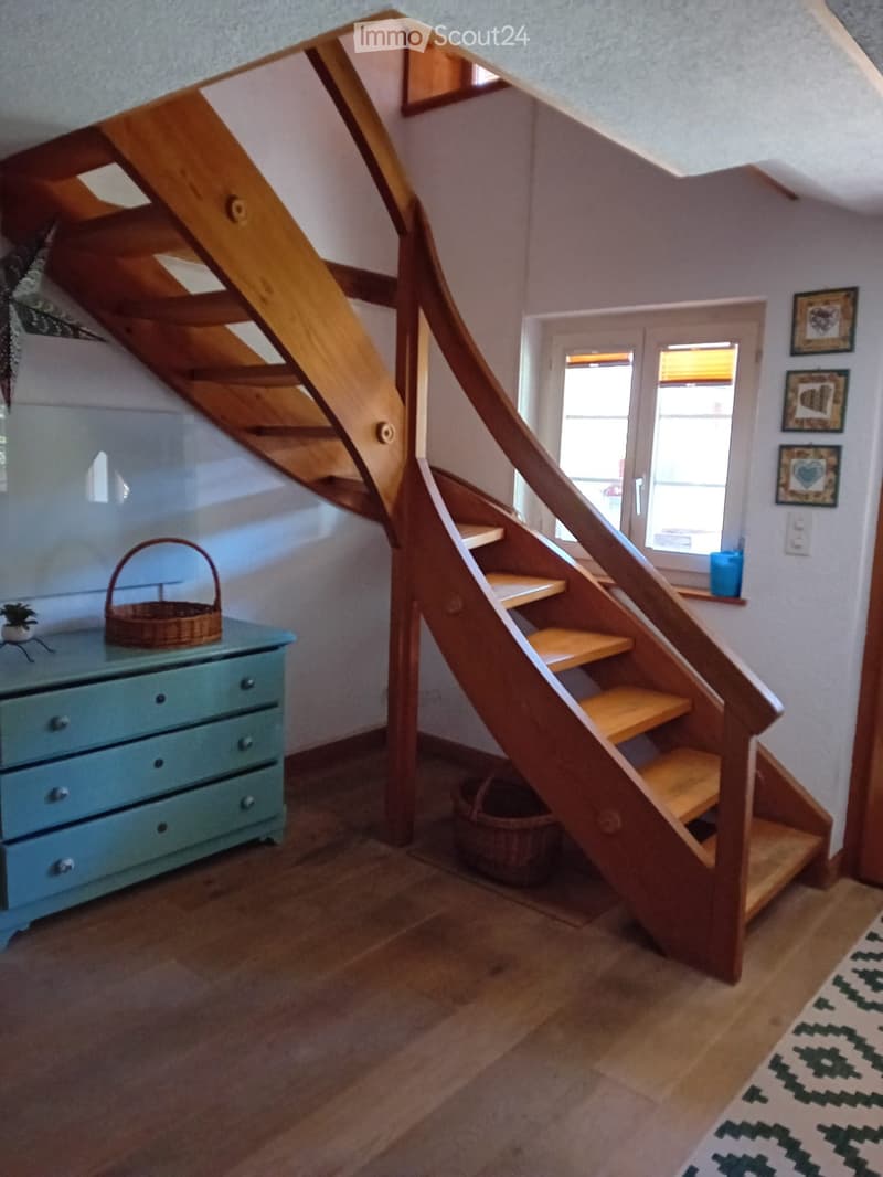 A wooden staircase with a modern design leads upwards, flanked by a light blue dresser with drawers, a small plant, and a woven basket on the landing. The hallway has wooden flooring and a patterned rug at the bottom.