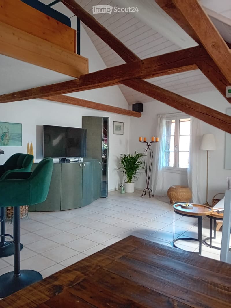Living room with exposed wooden beams, white tiled floor, green sofa, round wooden table, floor lamp, potted plant, and television on a cabinet.