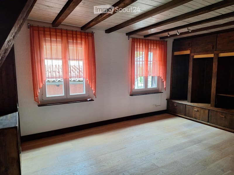 Empty room with wooden flooring, exposed wooden ceiling beams, built-in wooden cabinetry, and two windows with orange curtains.