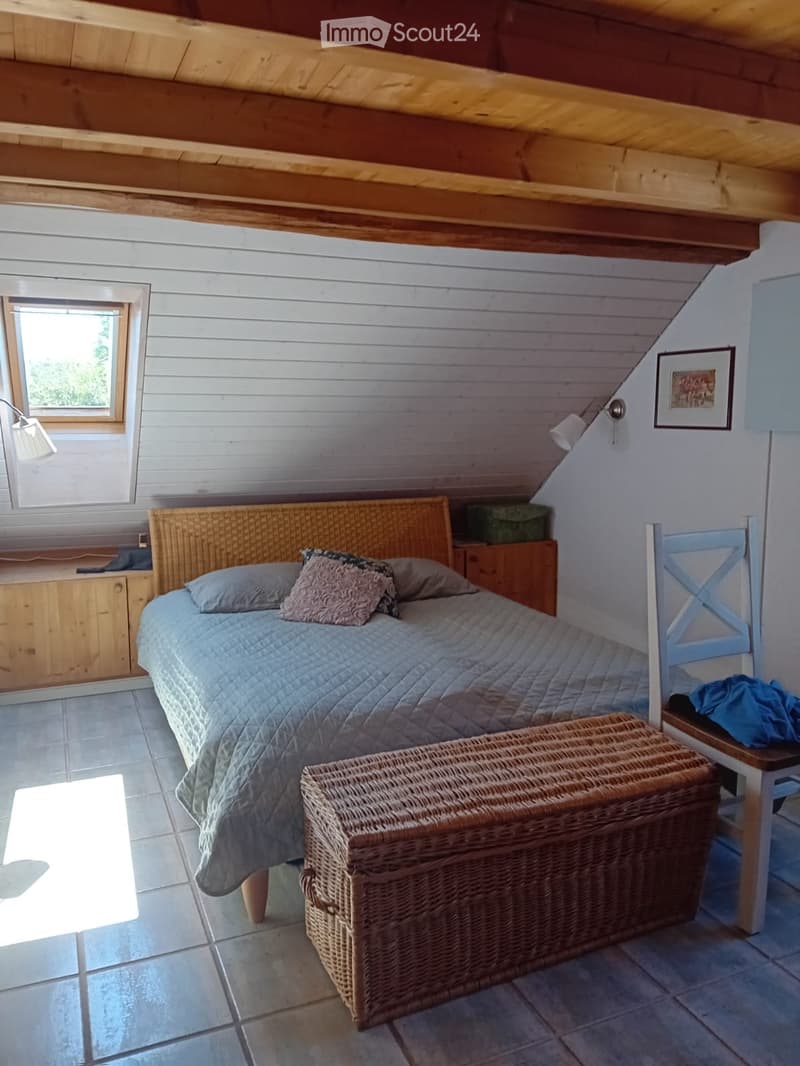 Cozy attic bedroom with slanted wooden ceiling, white paneled walls, tiled floor, and large window. Features a wicker headboard bed with pillows, a wicker storage box, a wooden chair, and a small cabinet.
