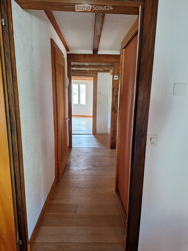 Wooden hallway with light-colored walls, wooden doors, and a visible room at the end with a window and wooden flooring.