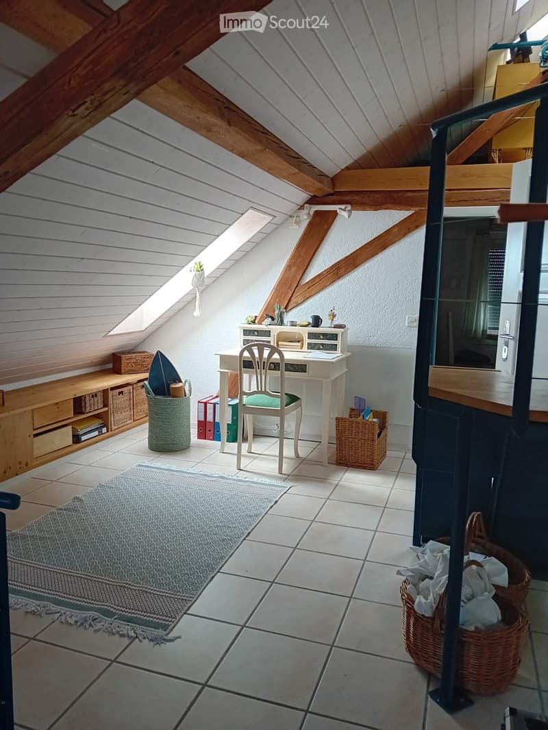 Cozy attic room with exposed wooden beams, skylight, tiled floor, and a small desk setup with a chair, wicker baskets, and a rug.